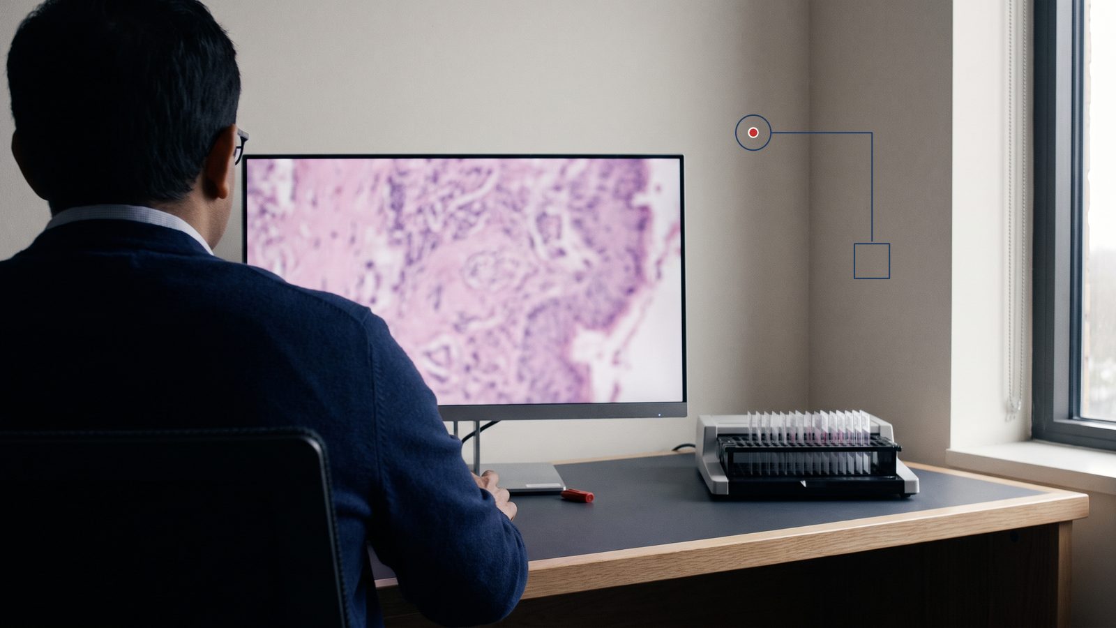 A pathologist reviews a stained tissue section on a large monitor, with a rack of glass slides on the desk beside him