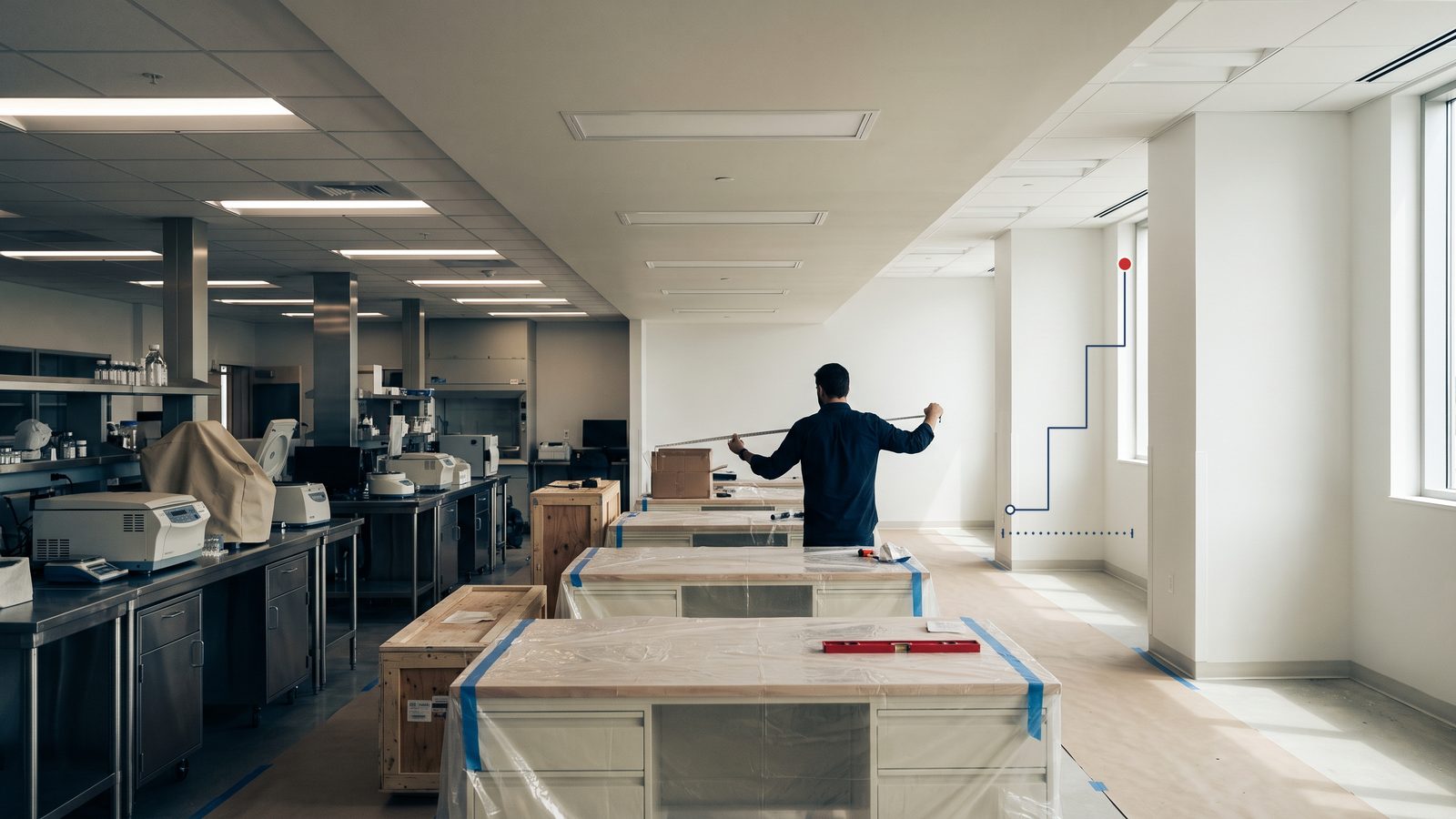 A man stretches a tape measure across an unfinished laboratory expansion, with new benches still wrapped in protective plastic
