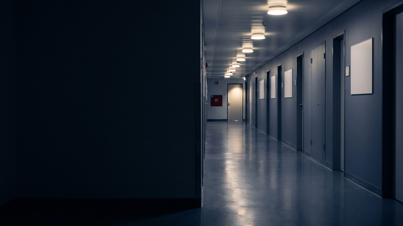 A long empty hospital corridor at night, closed doors along both walls and the floor reflecting the lights