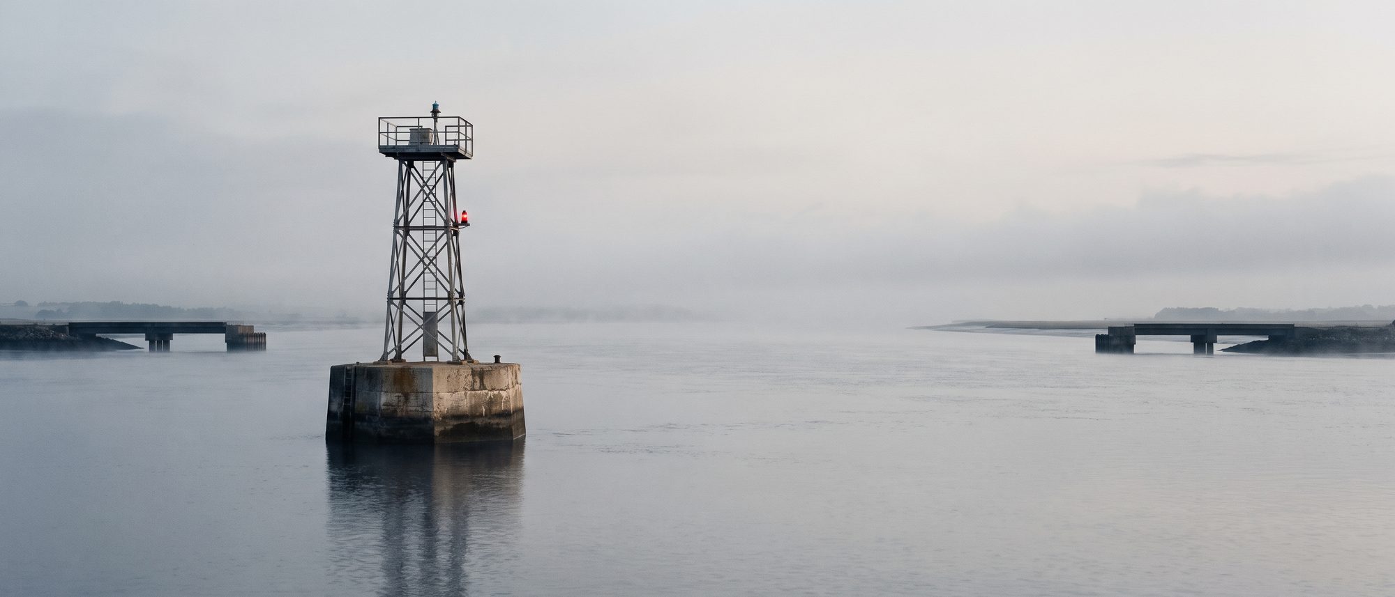 A single steel caisson tower standing alone in open water between two unfinished causeways at dawn.