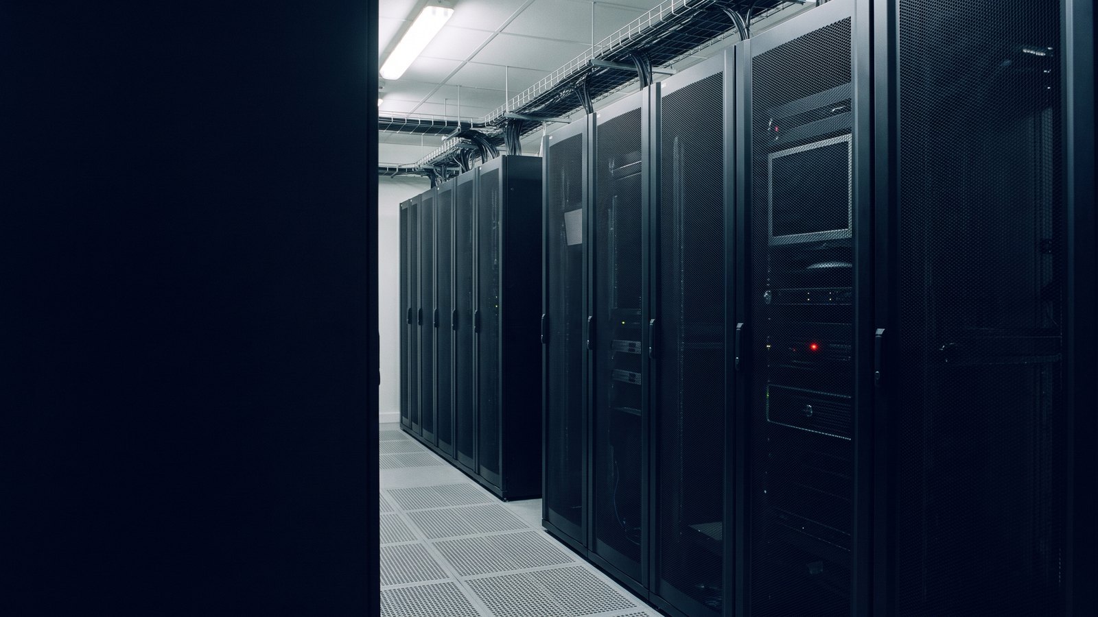 An aisle between two rows of black server racks in a data hall, with one small red status light lit